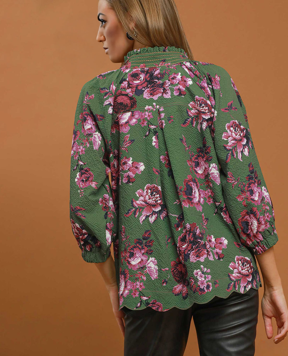 A woman shown from the back wearing a green blouse with a pink and purple floral print, paired with black leather pants, against a solid brown studio background.