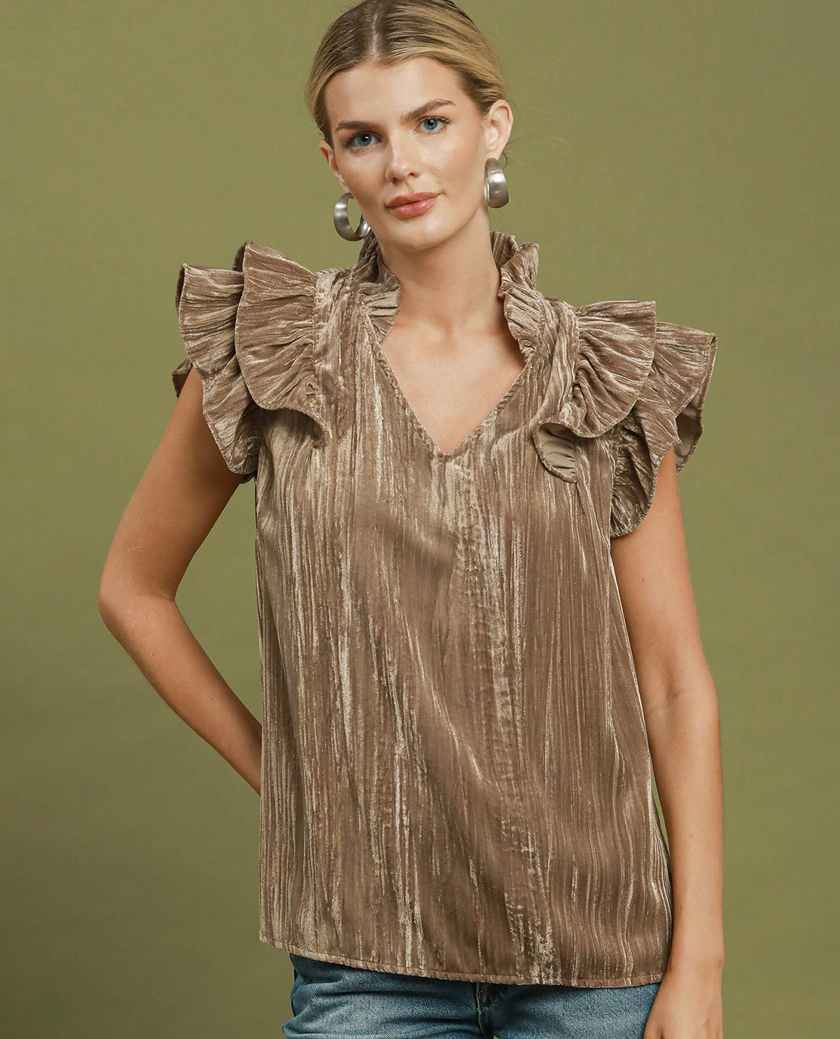 A woman wearing a sleeveless metallic taupe top with dramatic ruffled shoulders and a V-neckline, paired with blue jeans, set against a solid olive green studio background.