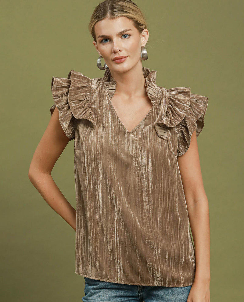 A woman wearing a sleeveless metallic taupe top with dramatic ruffled shoulders and a V-neckline, paired with blue jeans, set against a solid olive green studio background.