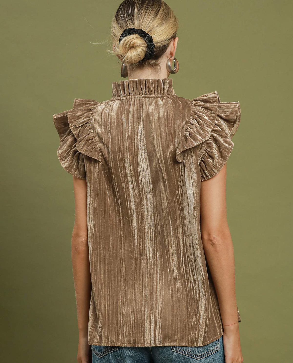 A woman viewed from the back wearing a taupe-brown ruffled velvet blouse with a high ruffled collar, set against a solid olive green background.