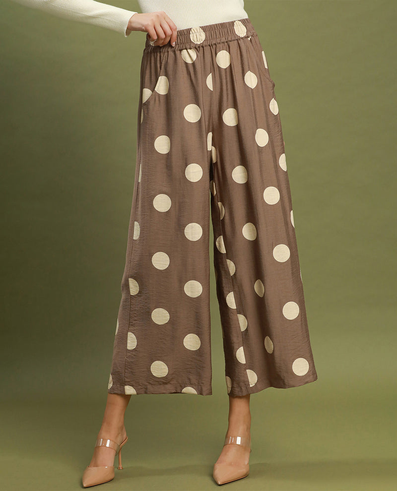 A model wears brown wide-leg cropped pants with large cream polka dots, paired with a white long-sleeve top and beige high heels, set against a solid olive green studio background.