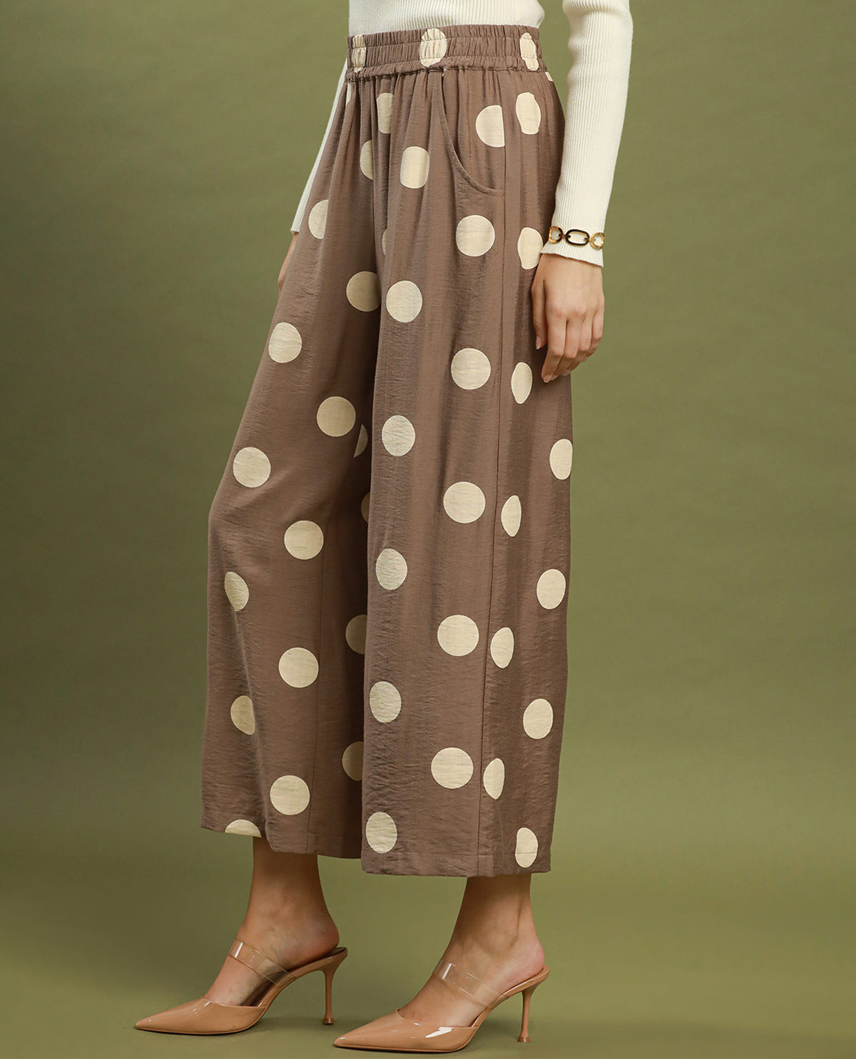 A model wearing brown wide-leg cropped pants with large cream polka dots, styled with a cream long-sleeve top and nude high-heeled mule sandals against an olive green studio background.