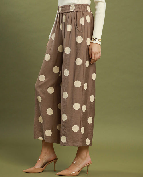 A model wearing brown wide-leg cropped pants with large cream polka dots, styled with a cream long-sleeve top and nude high-heeled mule sandals against an olive green studio background.