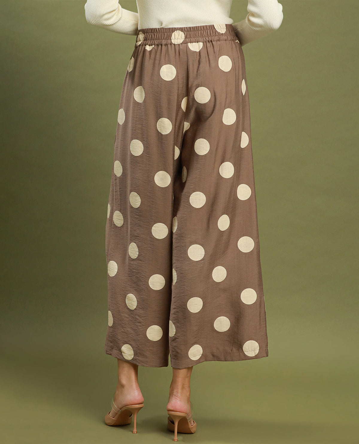 A person shown from the back wearing loose, wide-leg cropped pants in a taupe-brown color with large cream polka dots, paired with a white top and nude high-heel sandals, against a solid olive green studio background.