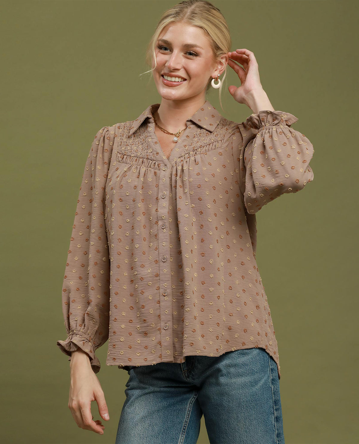 A woman modeling a brown long-sleeve blouse with a small geometric pattern, ruffled collar, and gathered puff sleeves, paired with blue jeans against an olive green background.