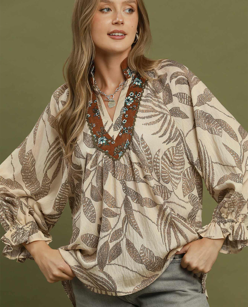 A woman wearing a beige blouse with a brown leaf pattern and a decorative brown embroidered neckline, posed against a solid olive green background.
