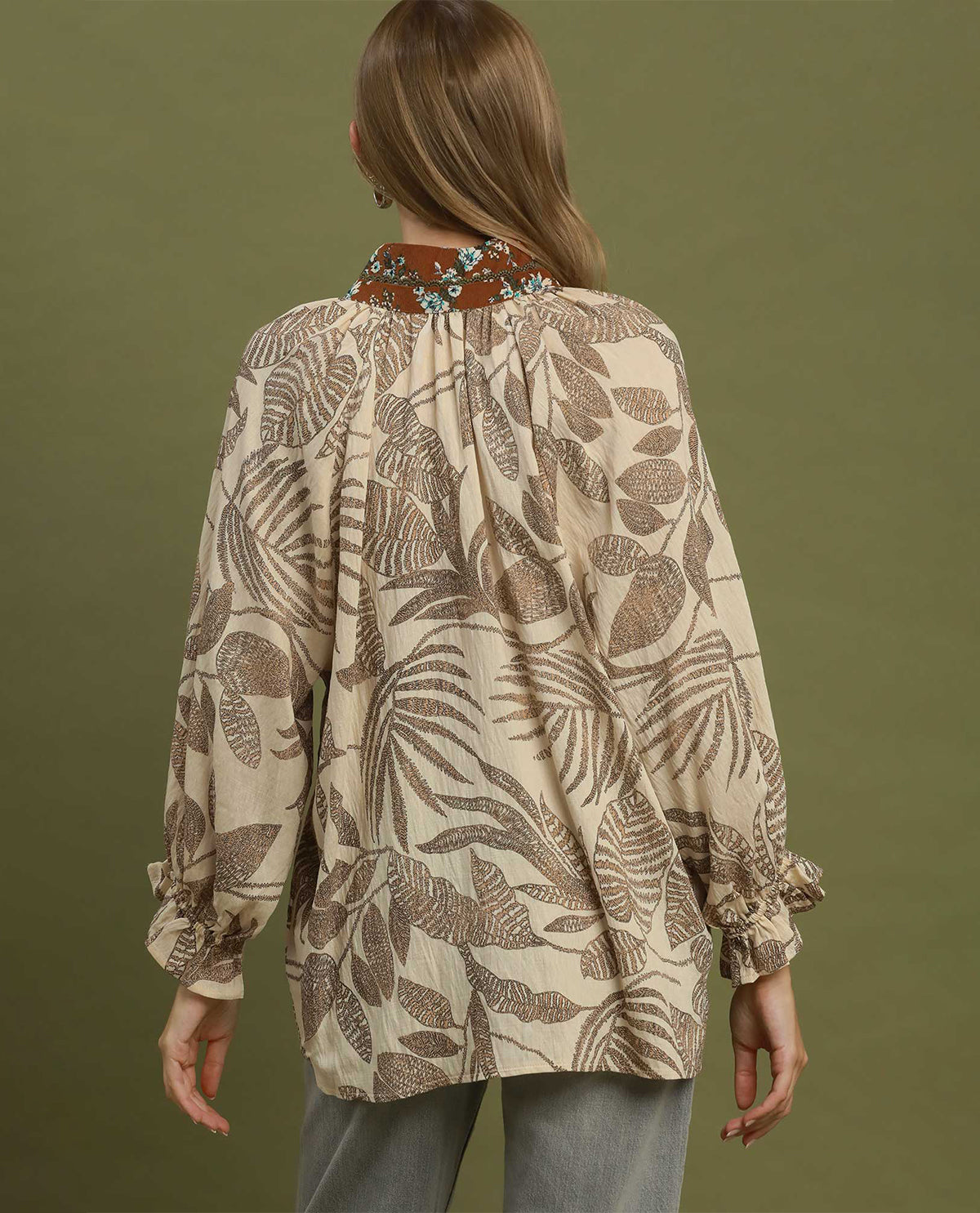 A woman viewed from the back wearing a beige blouse with a brown tropical leaf print, gathered neckline, and embroidered collar detail, paired with gray pants against an olive green background.