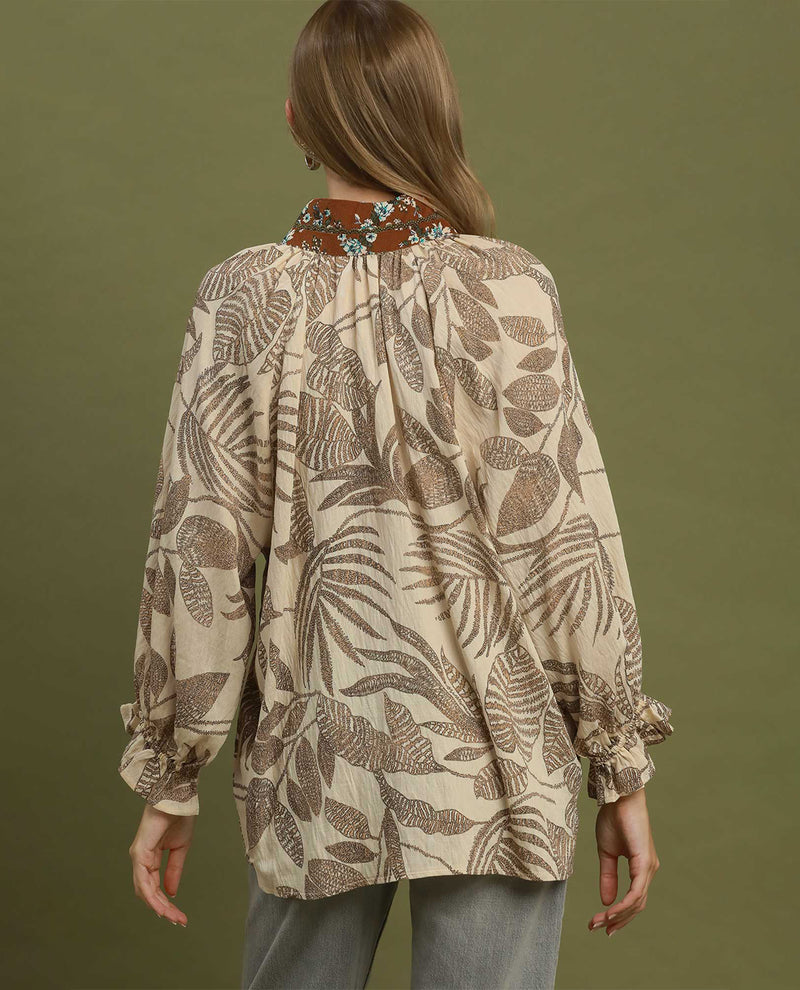 A woman viewed from the back wearing a beige blouse with a brown tropical leaf print, gathered neckline, and embroidered collar detail, paired with gray pants against an olive green background.