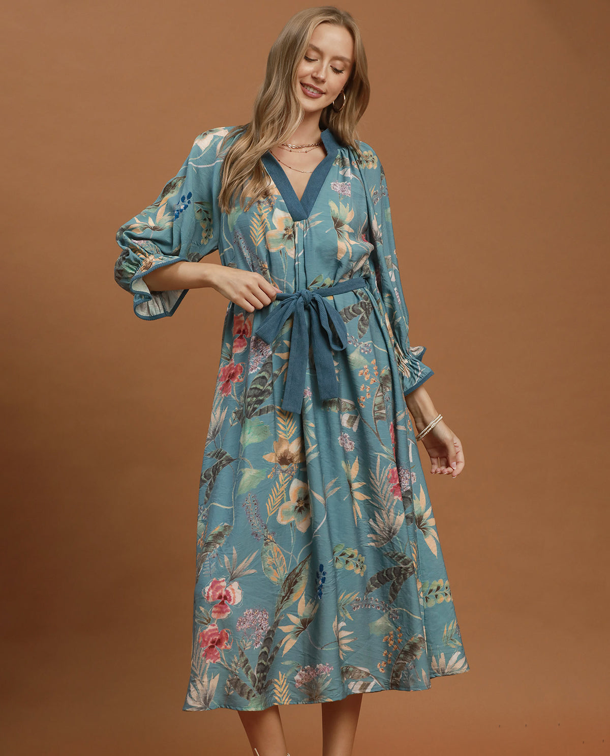 A woman modeling a teal blue floral midi dress with a V-neck, long sleeves, and a matching waist tie, set against a solid brown studio background.
