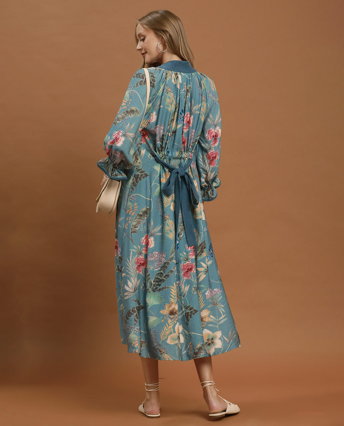 A woman modeling a long-sleeved midi dress with a blue base and a pink, red, and green floral print, shown from the back against a solid brown studio backdrop.