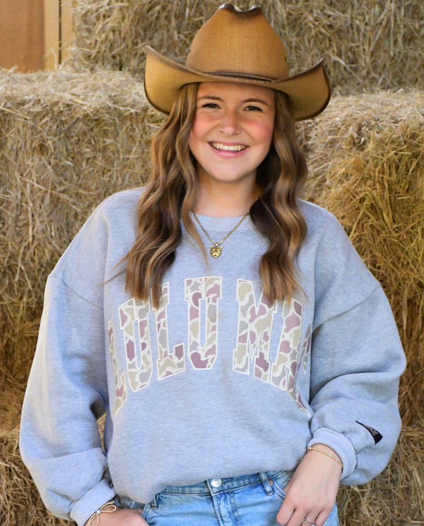 A smiling young woman wearing a brown cowboy hat and a light gray sweatshirt with a camouflage patterned 'LULU MAC' graphic, posed in front of a stack of hay bales.