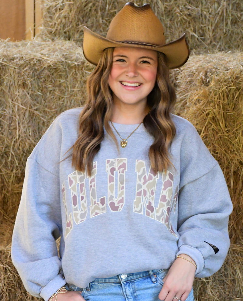 A smiling young woman wearing a brown cowboy hat and a light gray sweatshirt with a camouflage patterned 'LULU MAC' graphic, posed in front of a stack of hay bales.