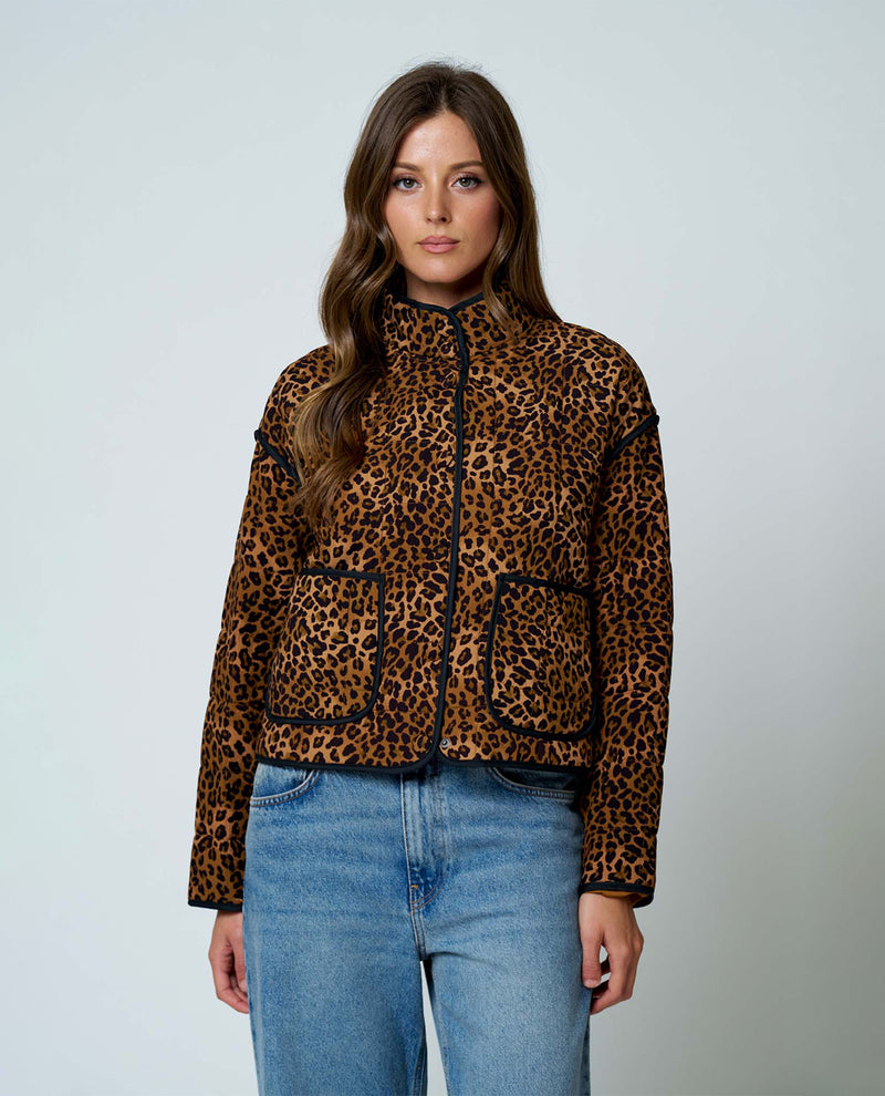 A woman modeling a leopard print jacket with black trim and front pockets, paired with blue jeans, against a light gray studio background.