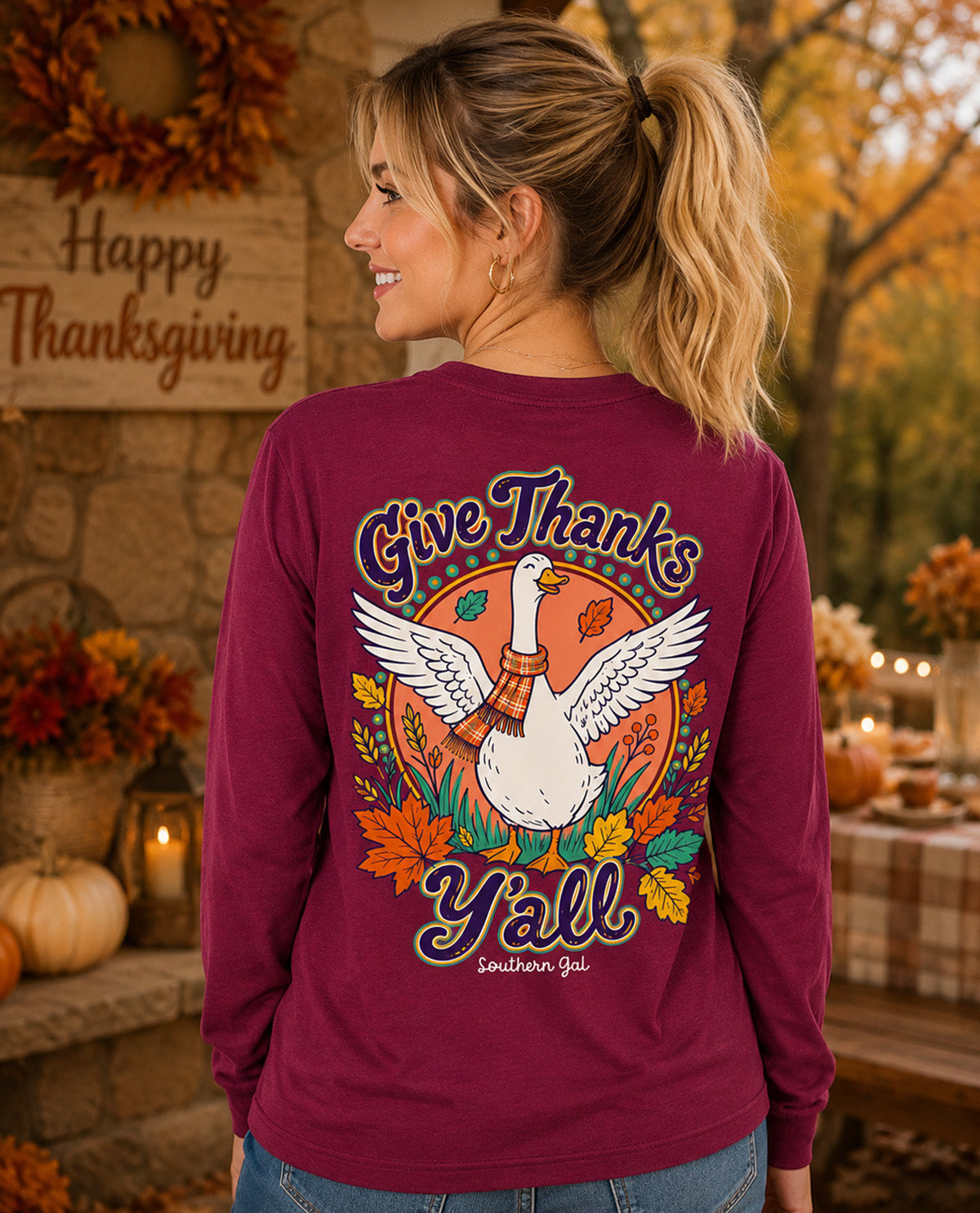 A woman shown from the back wearing a maroon long-sleeve shirt with a large Thanksgiving graphic featuring a white goose, autumn leaves, and the text 'Give Thanks Y'all' in a fall-themed setting.