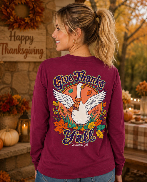 A woman shown from the back wearing a maroon long-sleeve shirt with a large Thanksgiving graphic featuring a white goose, autumn leaves, and the text 'Give Thanks Y'all' in a fall-themed setting.