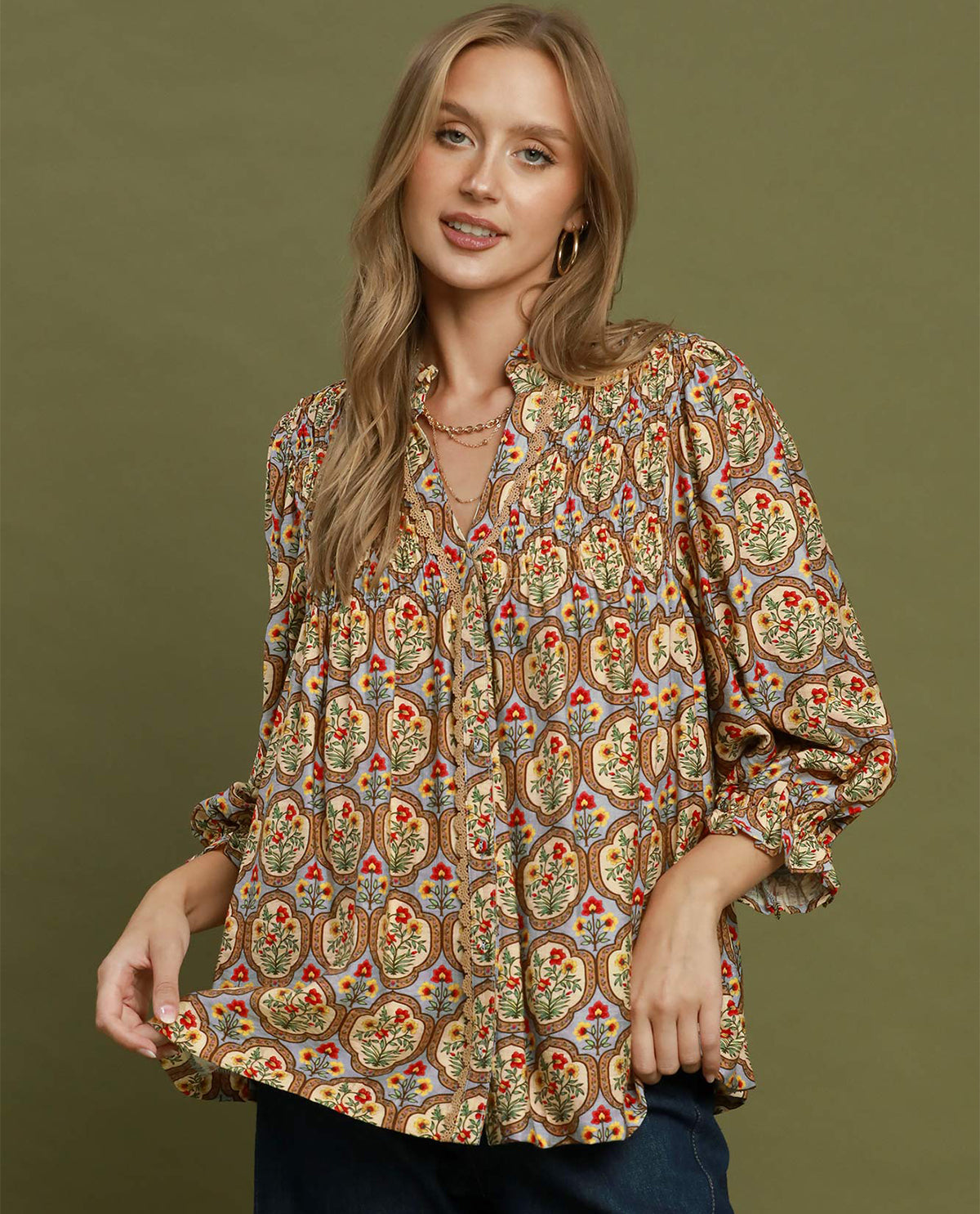 A woman wearing a long-sleeve bohemian-style blouse with a detailed multicolor paisley pattern, paired with dark blue jeans, set against a solid olive green background.