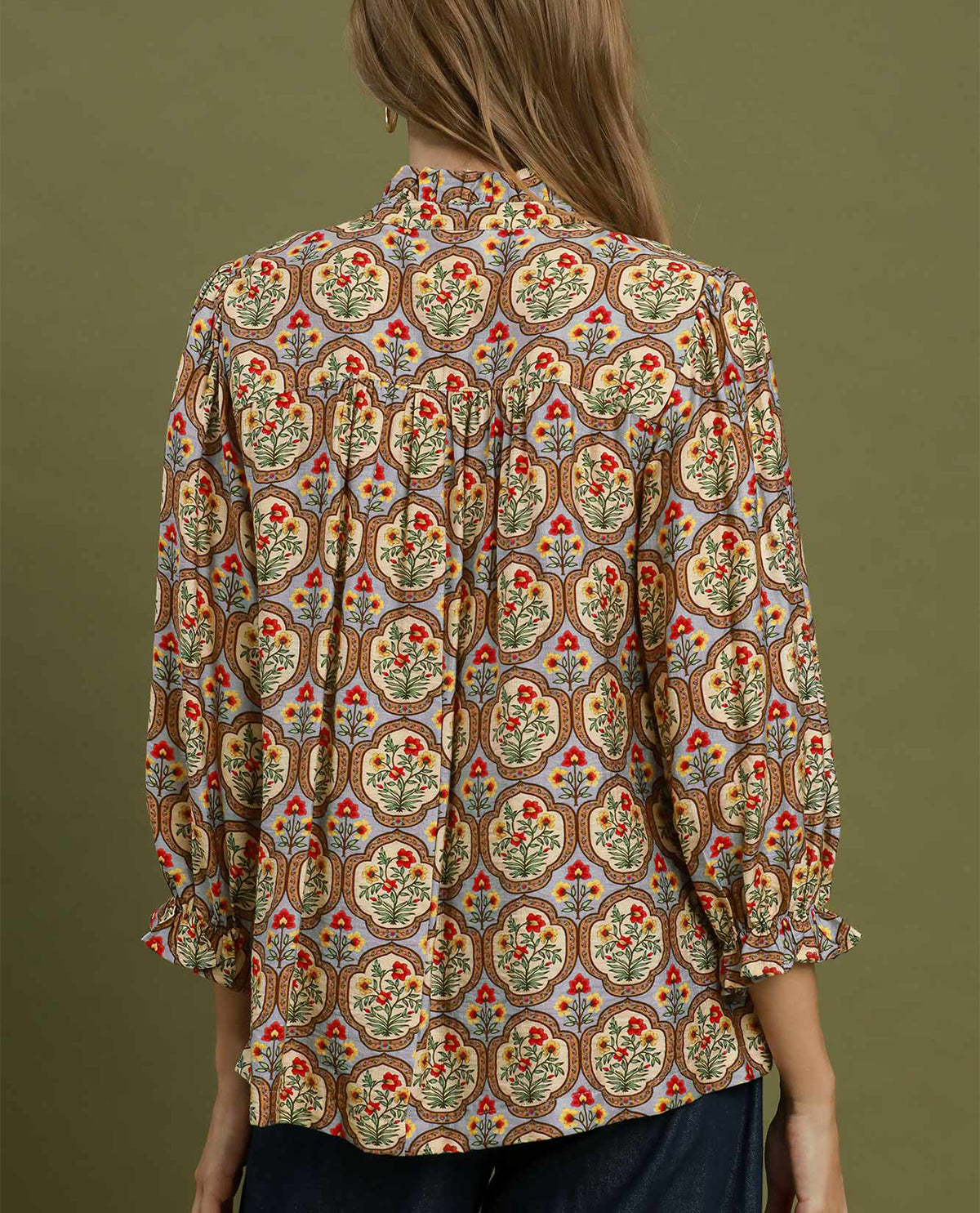 A woman shown from the back wearing a long-sleeve blouse with an intricate multicolor floral and geometric pattern on a beige base, set against a solid olive green background.