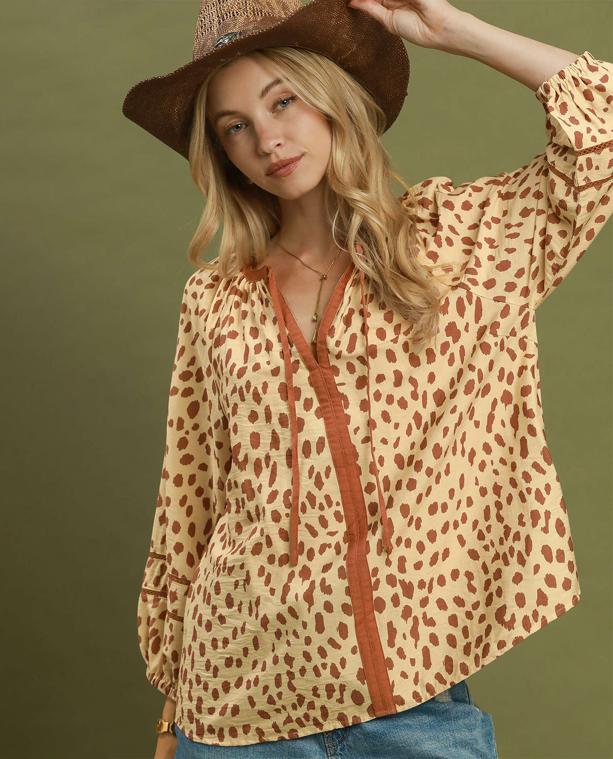 A woman wearing a beige long-sleeve blouse with a brown leopard print pattern, a brown faux-leather front panel with a drawstring, and a brown cowboy hat, posed against a solid olive green background.