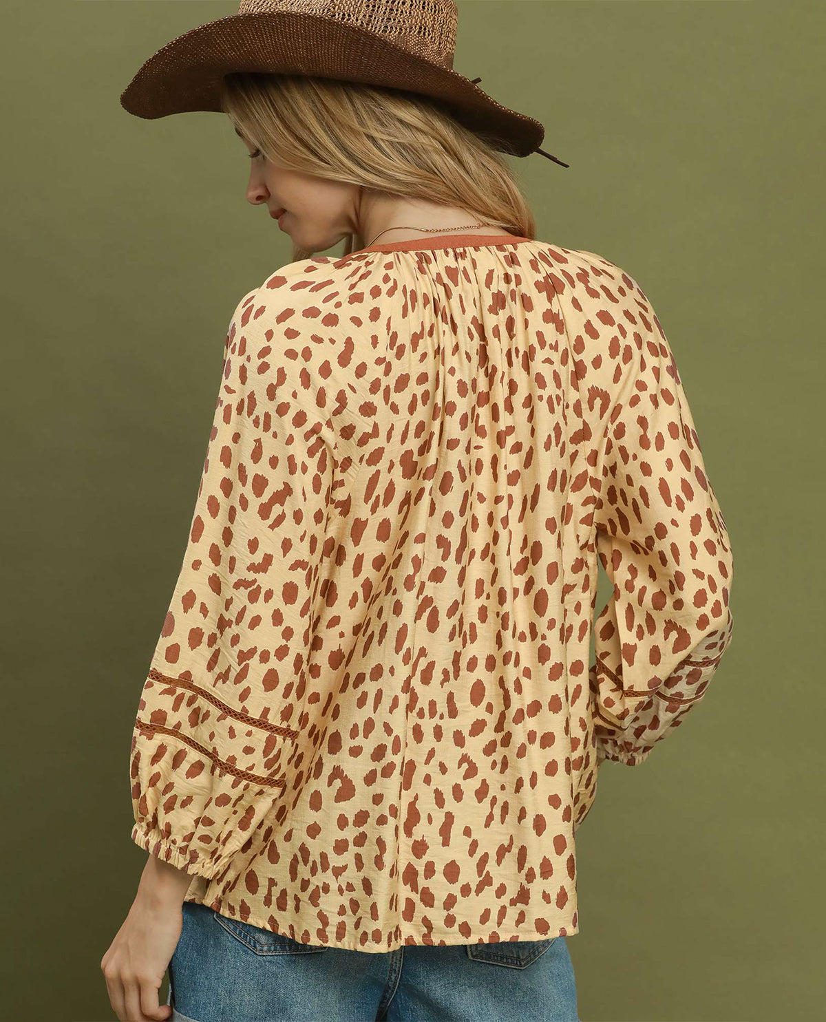 A woman wearing a beige blouse with a brown leopard print pattern, shown from the back. She is also wearing a brown cowboy hat and blue jeans, set against a solid olive green background.