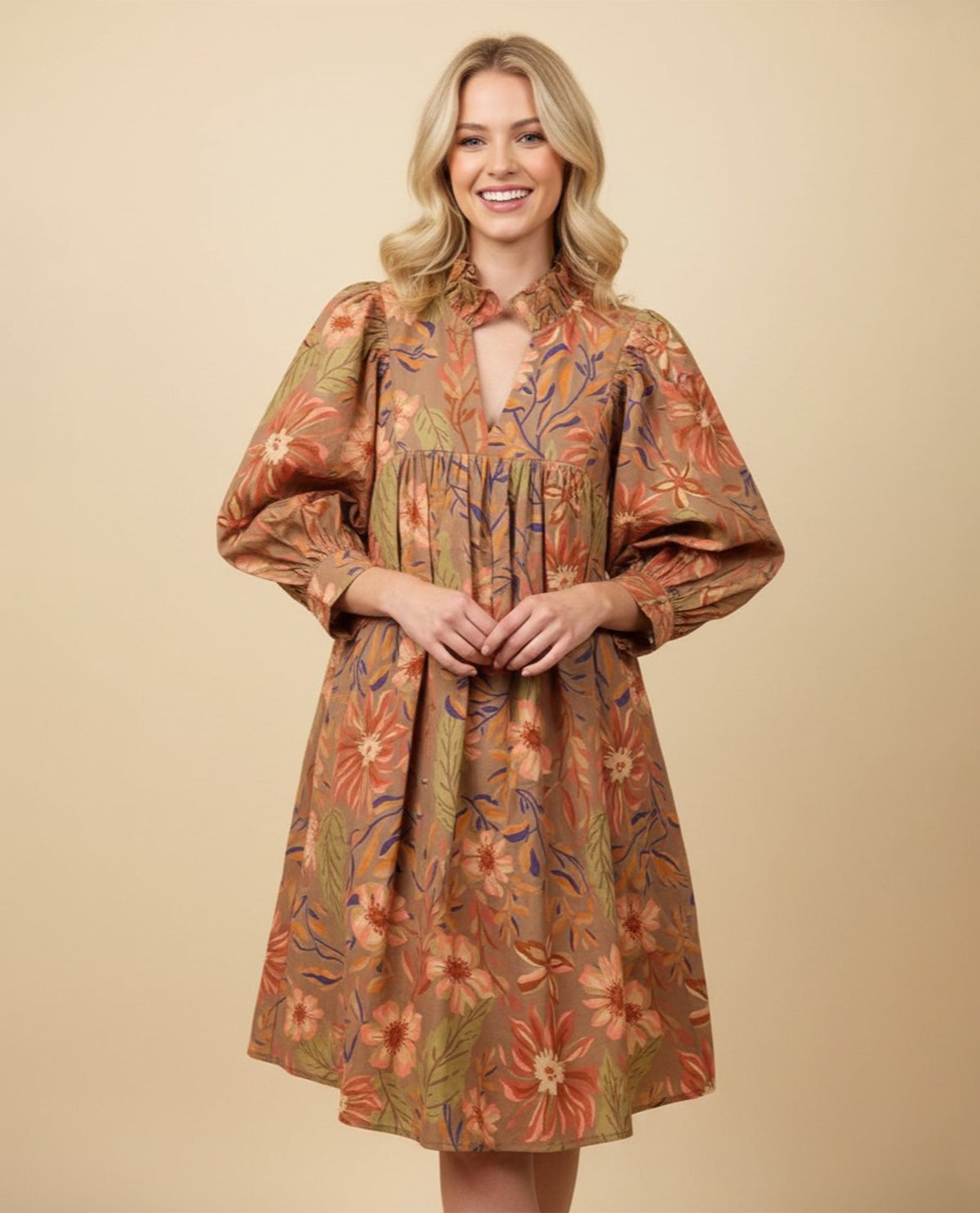 A woman modeling a knee-length floral dress with a high gathered neckline, long balloon sleeves, and an allover warm-toned print on a solid beige studio background.
