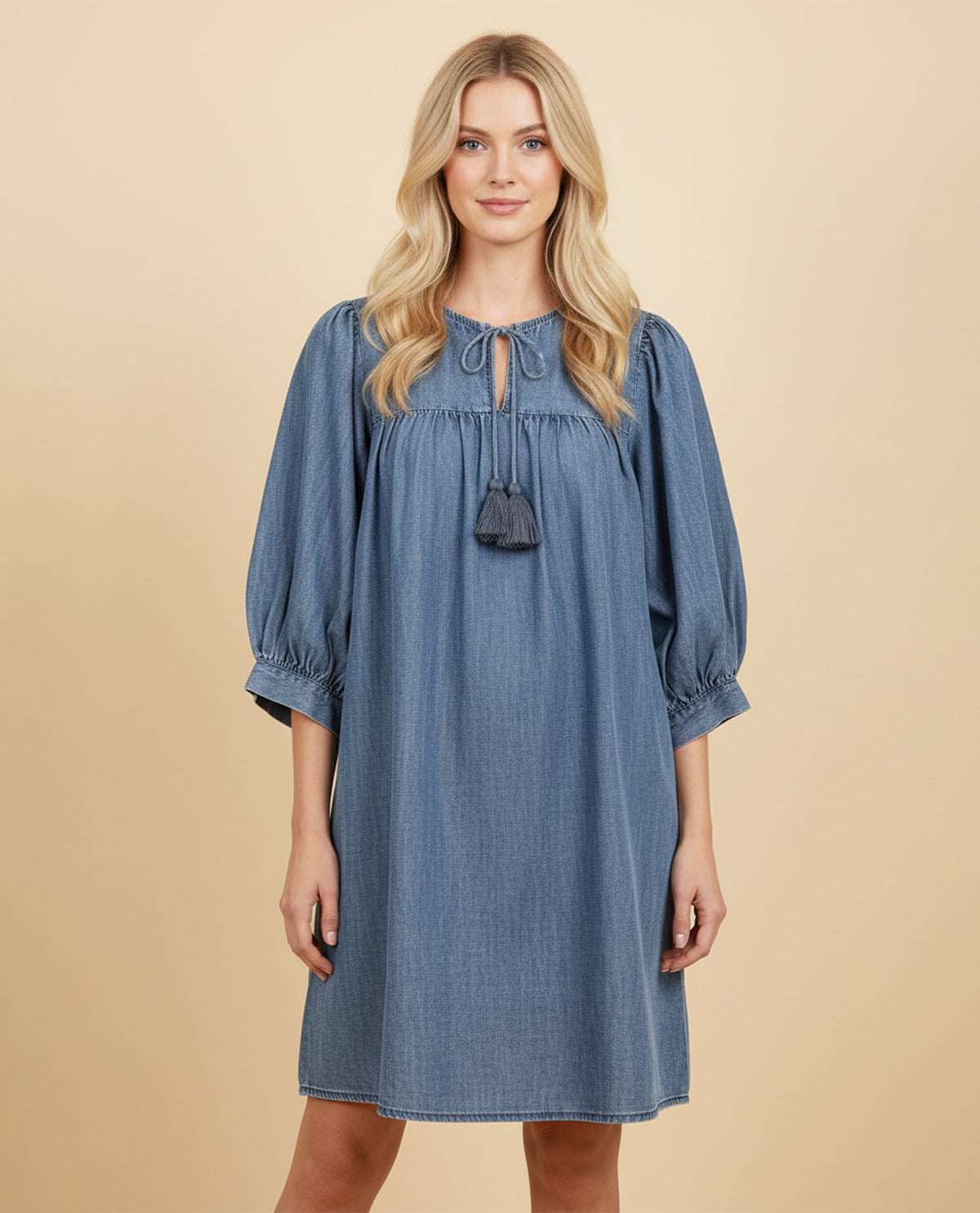 A woman modeling a blue denim-style dress with three-quarter puff sleeves, a tie neckline, and a small hanging tassel detail, set against a solid beige studio background.