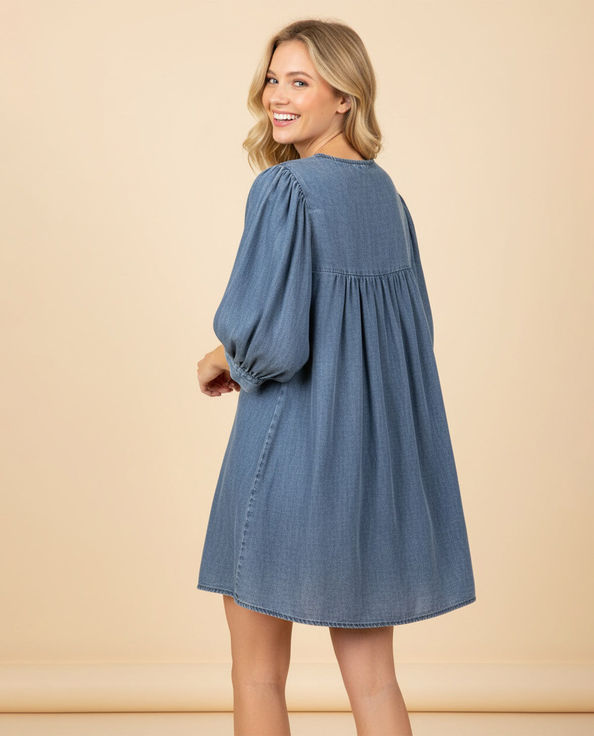 A woman wearing a blue denim-style mini dress with voluminous elbow-length sleeves, shown from the back against a plain beige studio backdrop.