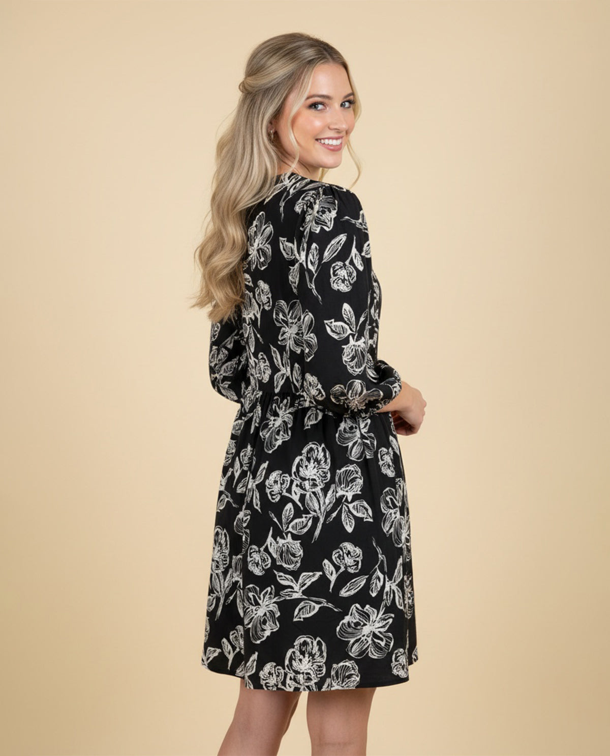 A woman modeling a black long-sleeve shift dress with a white floral print, photographed from the back against a solid beige studio background.