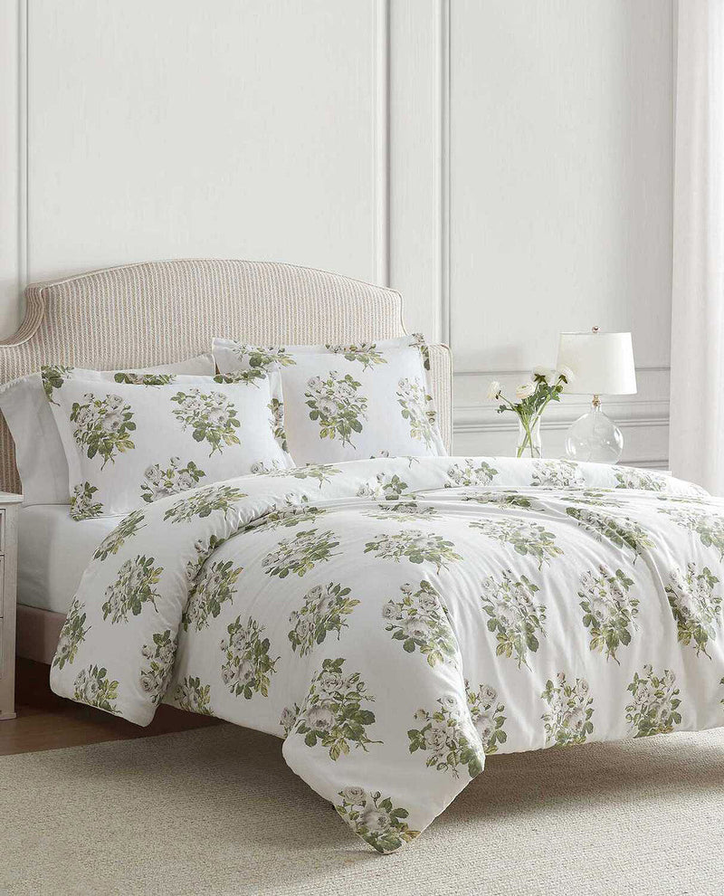 A white bedding set with a green floral and leaf pattern, styled on a bed with a beige upholstered headboard in a bright, neutral bedroom setting.