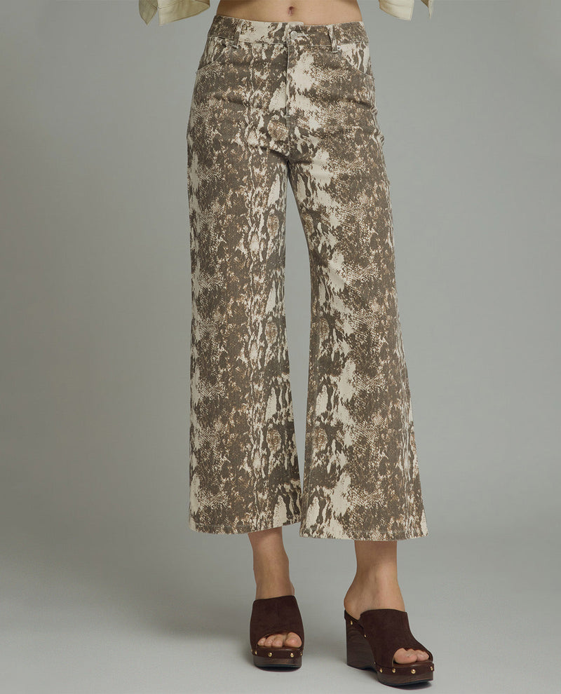 A person wearing cropped wide-leg pants with an abstract brown and cream print, paired with dark brown wedge sandals, photographed against a plain light gray studio background.