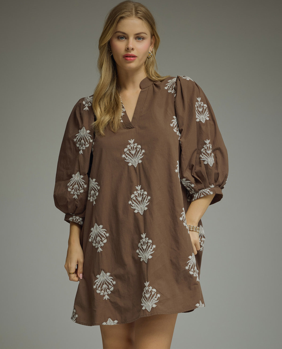 A woman modeling a brown tunic dress with white floral block prints, featuring a split neckline, voluminous puff sleeves, and a loose, comfortable fit against a solid gray studio background.
