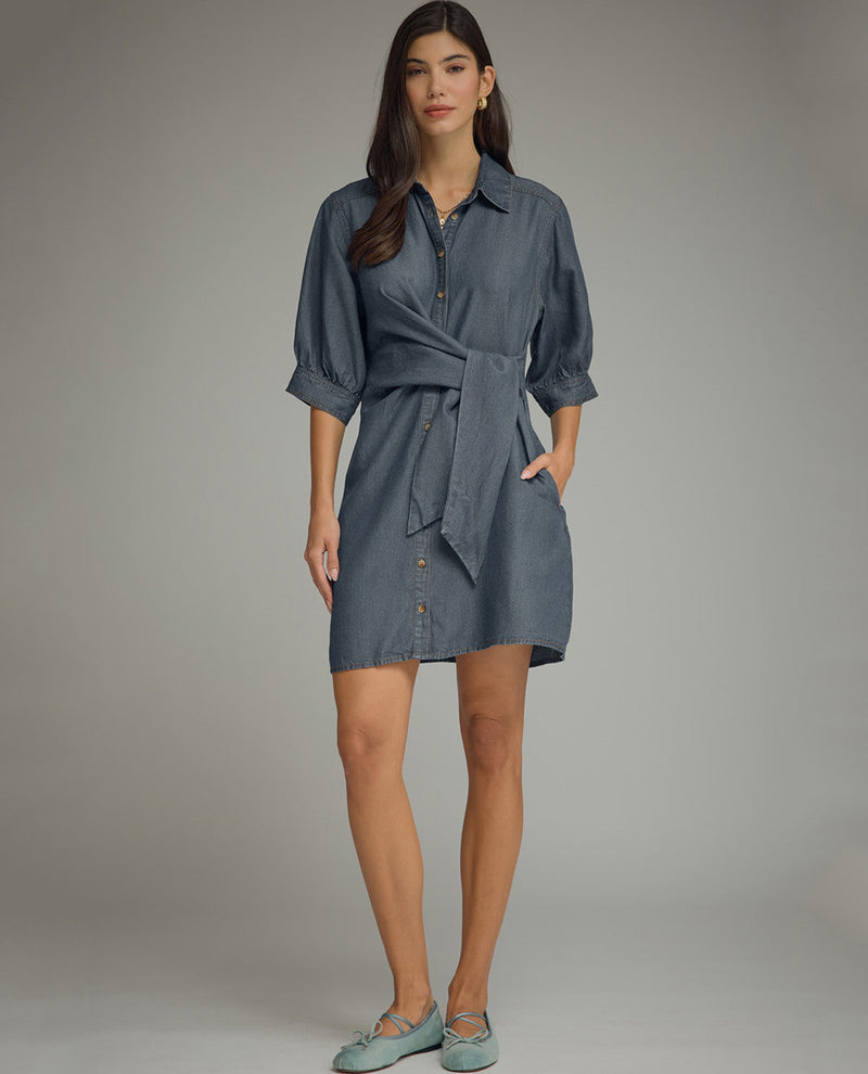 A woman modeling a blue-gray button-front shirt dress with a self-tie waist, three-quarter sleeves, and side pockets, paired with light blue flats, against a neutral gray studio background.