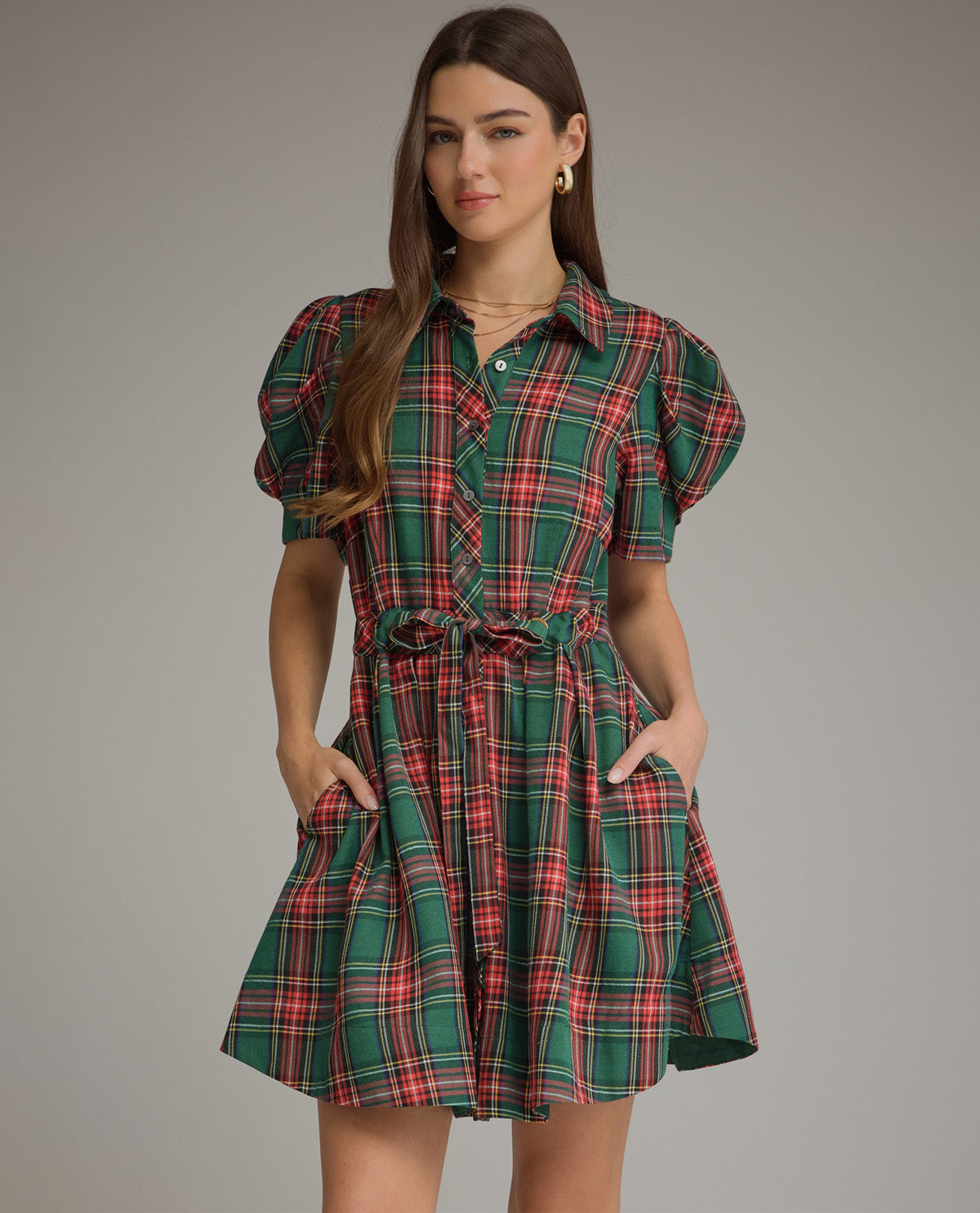 A woman modeling a green tartan plaid mini dress with puff sleeves, a button-down front, and a tie waist, set against a solid gray studio background.