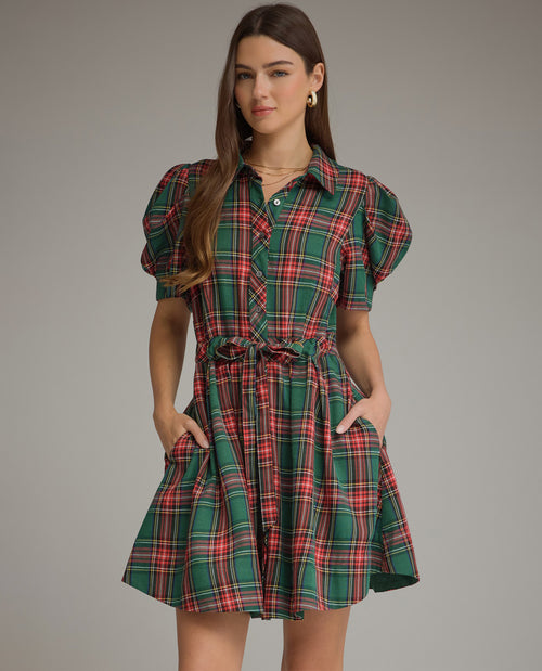 A woman modeling a green tartan plaid mini dress with puff sleeves, a button-down front, and a tie waist, set against a solid gray studio background.