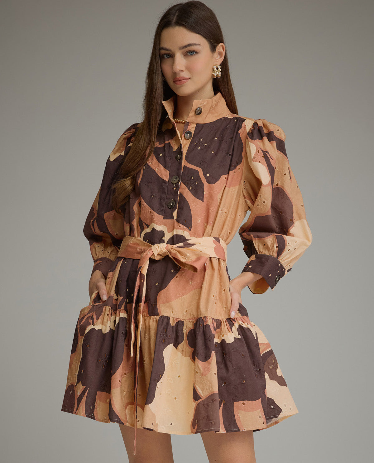 A woman models a short puff-sleeve shirt dress with a brown and beige abstract print, featuring a button-up front, a matching fabric tie belt, and a tiered ruffled hem, set against a solid gray studio background.