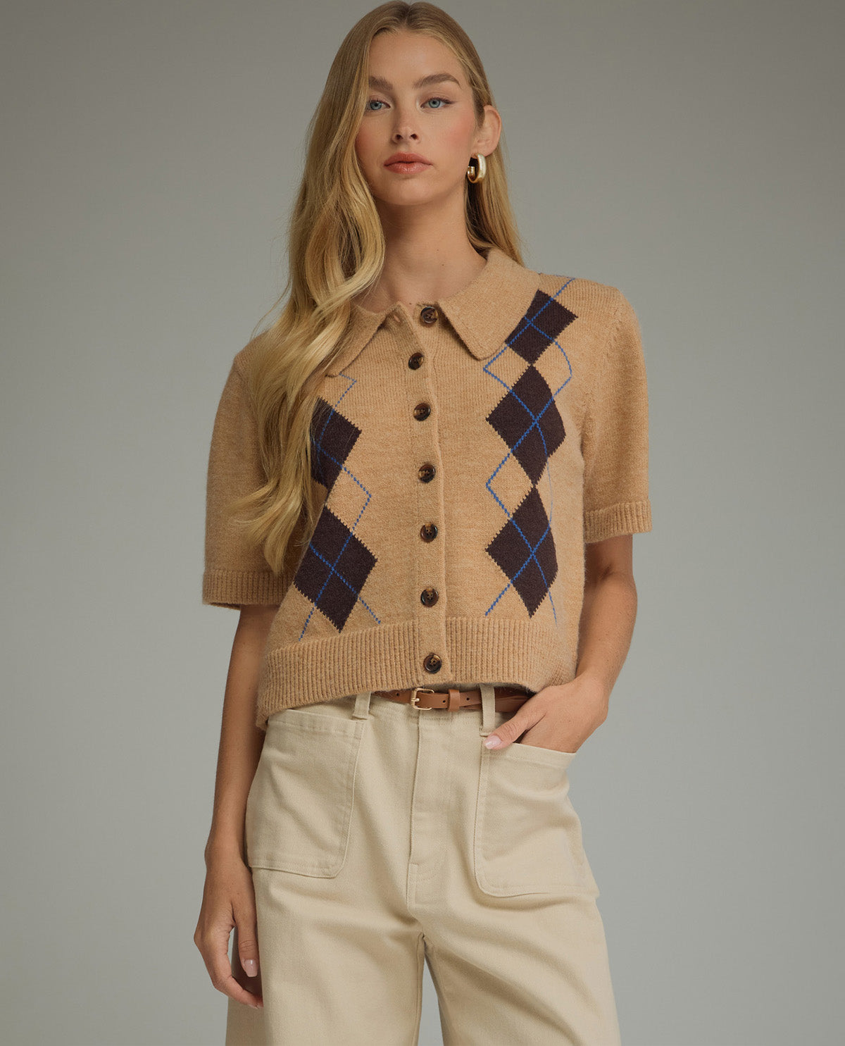 A woman modeling a short-sleeve knit button-front cardigan in camel beige with dark navy and blue argyle diamond patterns, paired with light beige pants against a plain gray studio background.