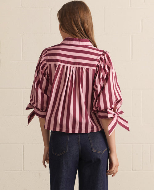 A woman viewed from the back wearing a burgundy and pink vertically striped blouse with pleated detailing and tie details on the sleeves, paired with dark blue jeans, set against a light beige brick wall.