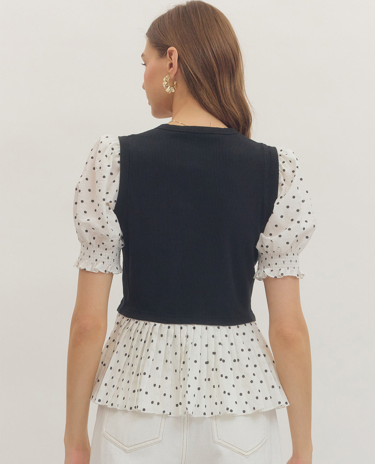 Woman wearing a black vest over a polka dot top with white pants on a plain background