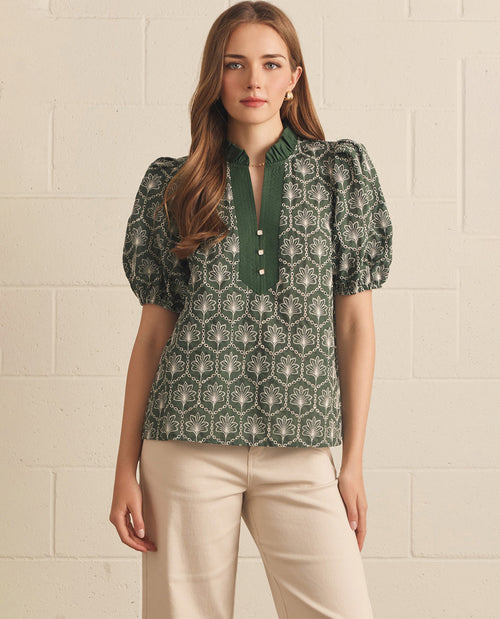 Woman wearing a green patterned blouse with beige pants against a beige brick wall.