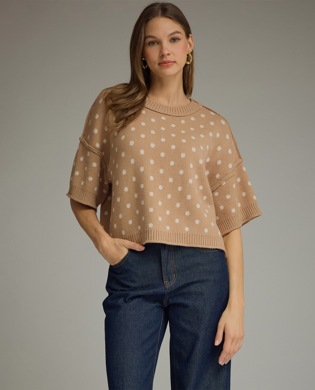 Woman wearing a beige polka dot sweater and blue jeans against a gray background