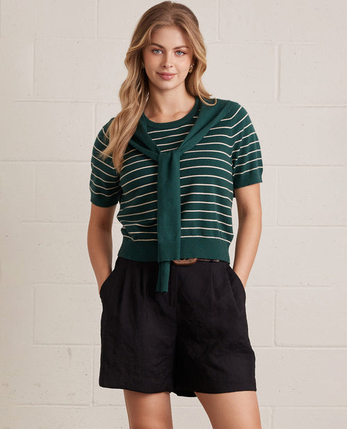 Woman wearing a green striped sweater and black shorts against a white brick wall.