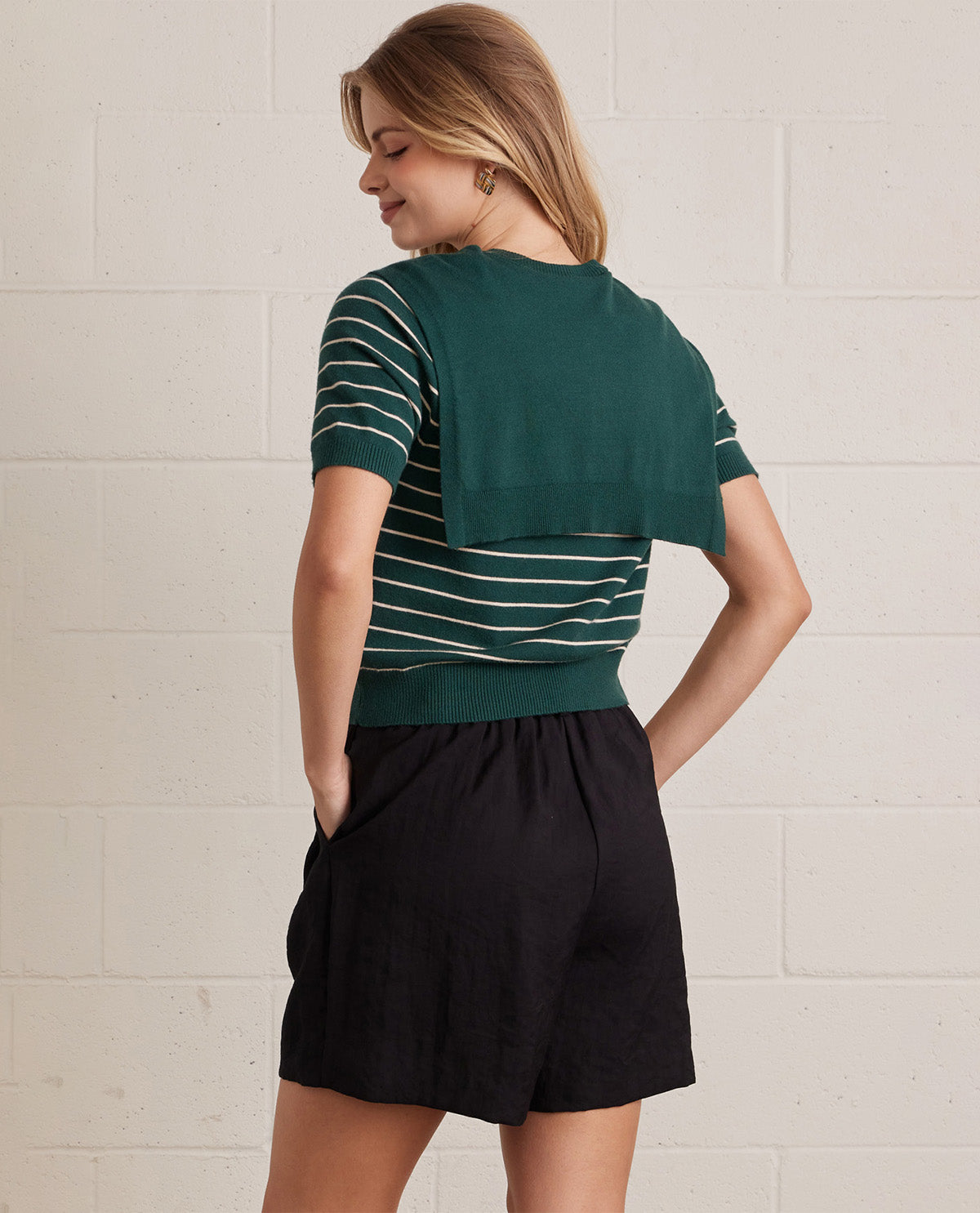 A woman wearing a dark green short-sleeve knit top with thin white horizontal stripes and a solid green draped cape-style back panel, paired with black shorts, shown from a rear three-quarter view against a white brick wall.