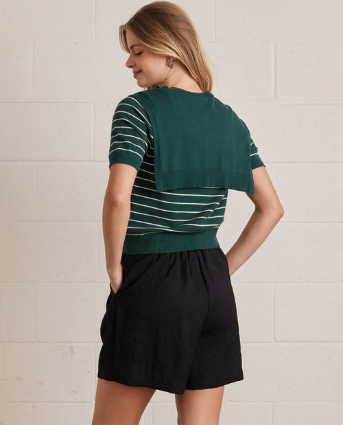 A woman wearing a dark green short-sleeve knit top with thin white horizontal stripes and a solid green draped cape-style back panel, paired with black shorts, shown from a rear three-quarter view against a white brick wall.