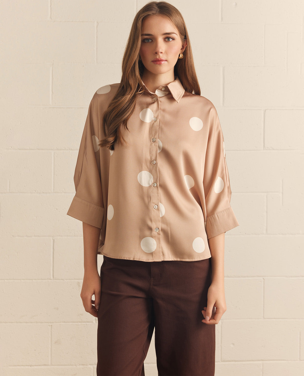 Woman wearing a beige blouse with white polka dots and brown pants against a beige tiled wall.