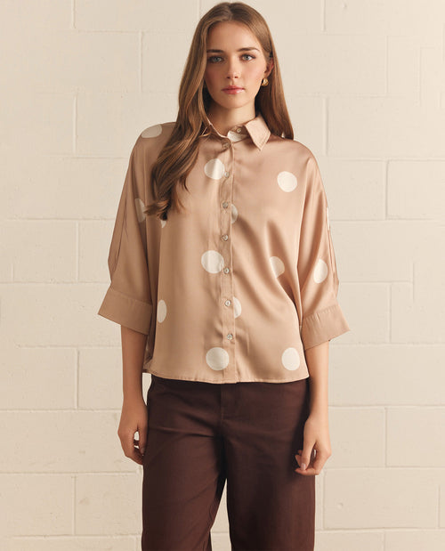 Woman wearing a beige blouse with white polka dots and brown pants against a beige tiled wall.