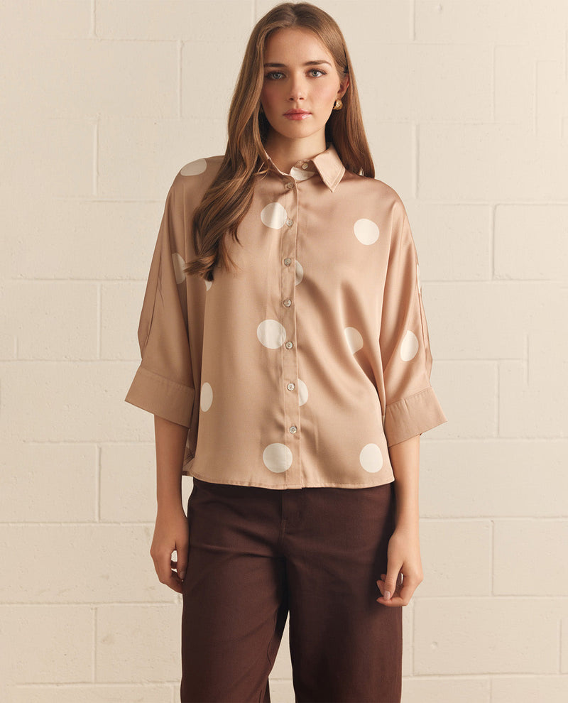 Woman wearing a beige blouse with white polka dots and brown pants against a beige tiled wall.