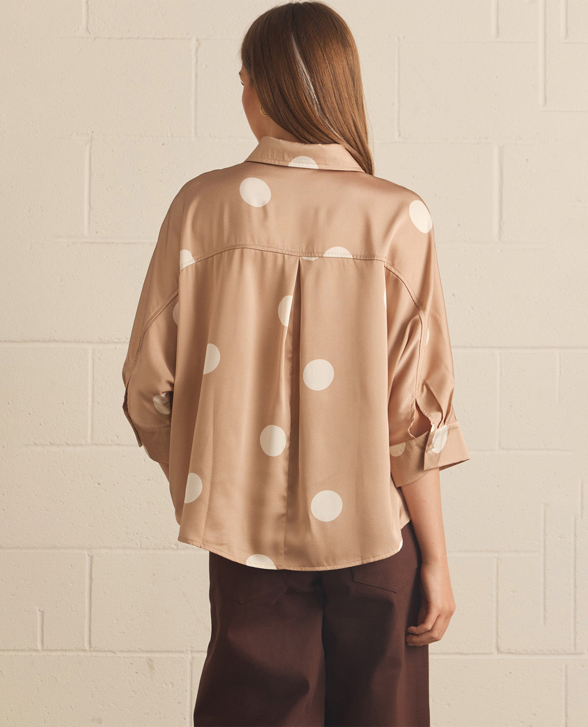 A woman viewed from behind wearing a beige satin button-up shirt with large white polka dots and dark brown trousers, standing against a white painted brick wall.