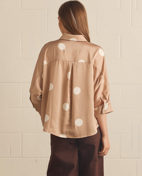 A woman viewed from behind wearing a beige satin button-up shirt with large white polka dots and dark brown trousers, standing against a white painted brick wall.