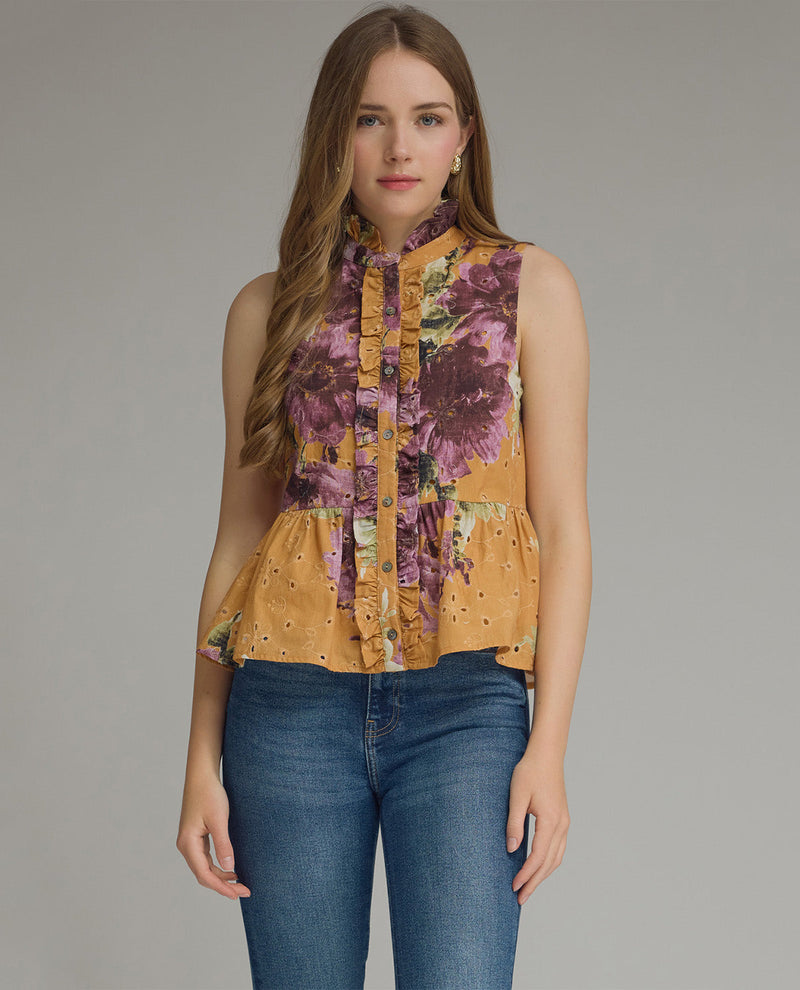 A woman modeling a sleeveless high-neck blouse with a ruffled front and a yellow floral print, paired with blue jeans, against a solid gray studio background.