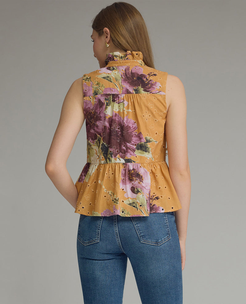 A woman shown from the back wearing a sleeveless high-neck peplum top in mustard yellow with a large floral print in purple, pink, and green, paired with blue jeans, against a solid gray studio background.