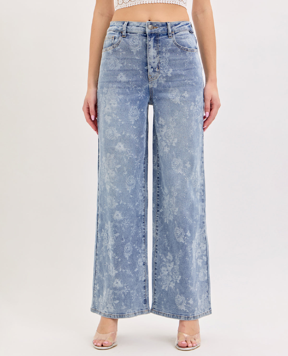A model wearing light blue wide-leg denim jeans with a faded floral pattern, paired with clear high-heeled sandals against a plain white studio background.